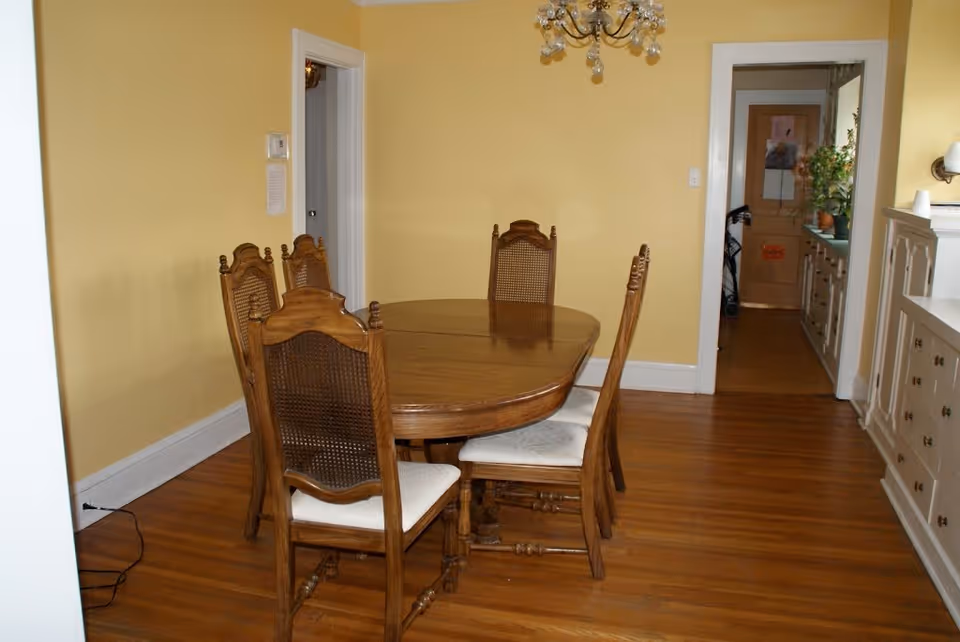 A dining room with yellow walls and wooden flooring featuring a wooden oval dining table surrounded by six wooden chairs with white cushions. A chandelier hangs above the table. There is a doorway leading to another room with cabinets and plants visible.