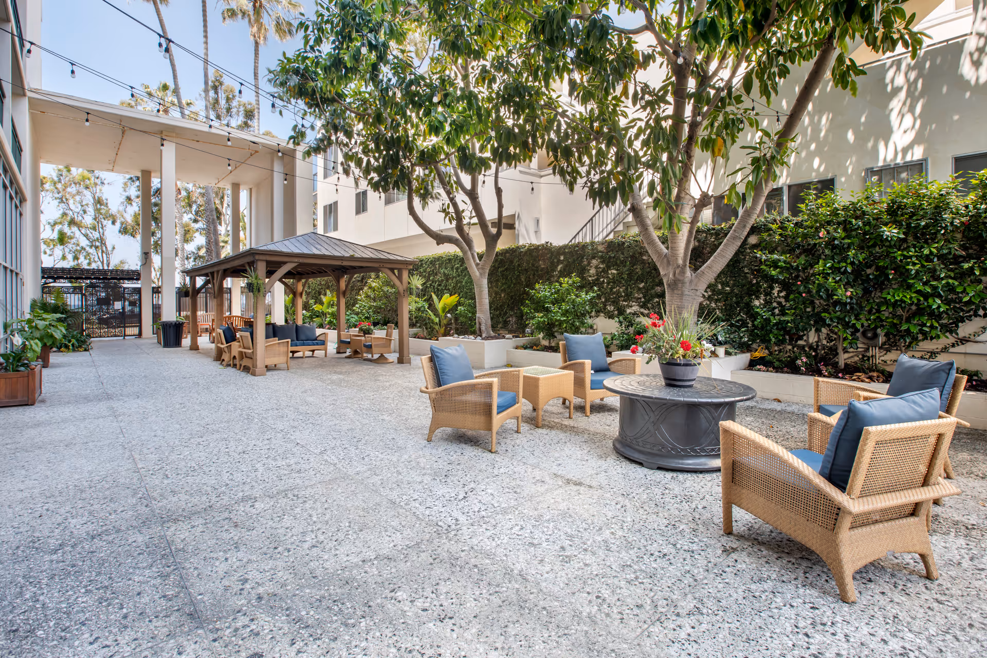 Outdoor patio area at Brookdale Ocean House featuring wicker chairs with blue cushions arranged around a circular fire pit table. There is a wooden gazebo with additional seating under string lights, surrounded by trees and greenery.