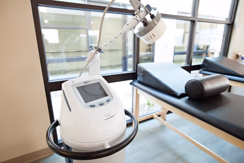 Physical therapy treatment room with a medical therapy device in front of padded treatment tables by large windows.