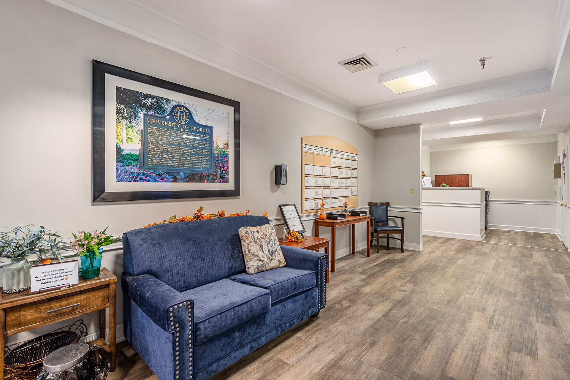 Interior view of a senior living facility lobby area with a blue upholstered couch, a wooden side table with plants and a sign, a framed University of Georgia plaque on the wall, a bulletin board, and a reception desk in the background.