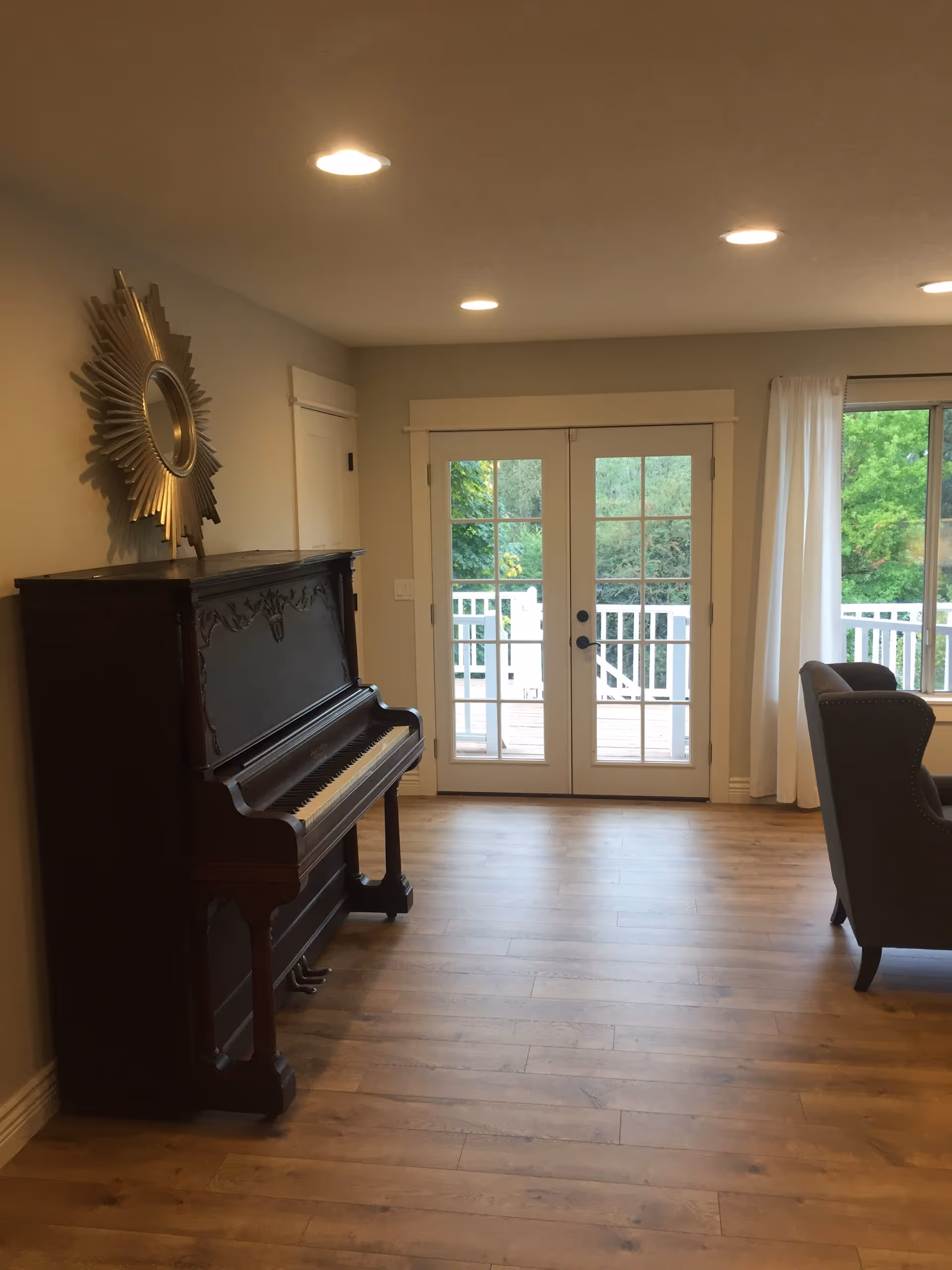 Bright living room with an upright piano against the left wall, French doors to a deck, a single armchair, and wood flooring.