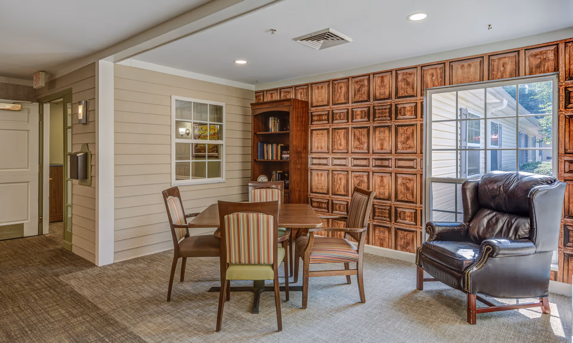 A cozy interior room with a wooden table surrounded by four chairs, two with striped upholstery and two with solid brown seats. There is a large leather armchair near a window that shows an exterior view of a white building. The walls have a decorative wooden panel design and a built-in bookshelf with books. The floor is carpeted and the ceiling has recessed lighting.