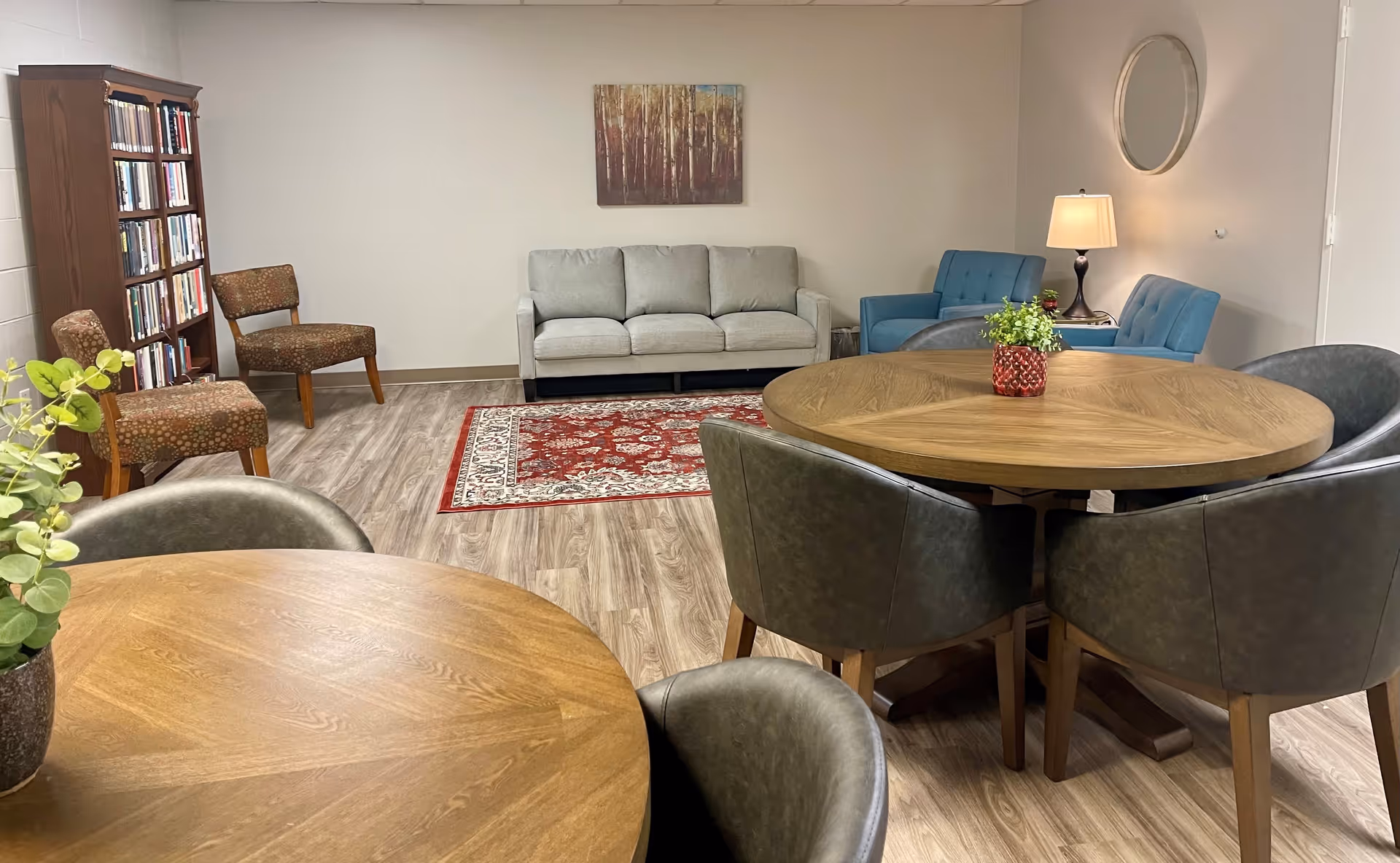 A cozy senior living common area with two round wooden tables surrounded by dark cushioned chairs, a gray sofa, two blue armchairs, a red patterned rug, a wooden bookshelf filled with books, two patterned chairs, a table lamp, a round wall mirror, and a painting of trees on a beige wall.
