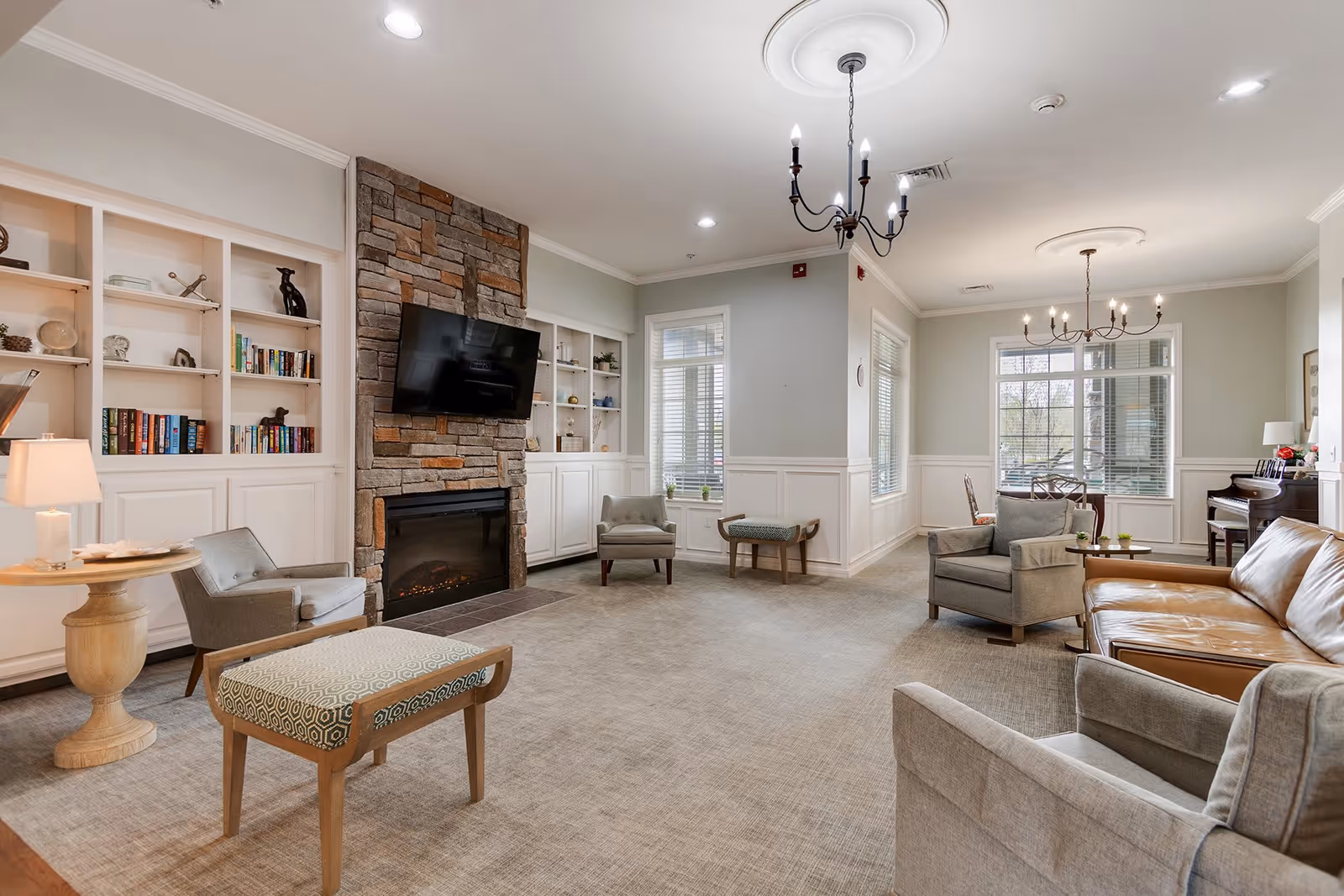 A spacious and well-lit living room with a stone fireplace and a mounted flat-screen TV. The room features built-in white shelves with books and decorative items, several armchairs, a leather sofa, a small wooden table with a lamp, and a piano near large windows with blinds. Two chandeliers hang from the ceiling, and the carpeted floor adds warmth to the space.