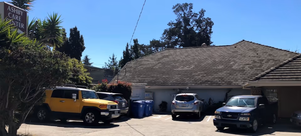 Parking lot area outside a single-story building with a shingled roof under a clear blue sky. Several cars are parked, including a yellow SUV, a silver car, a white car, and a blue truck. There is a sign on the left side that reads 'Ashby Care Center' surrounded by greenery and trees.