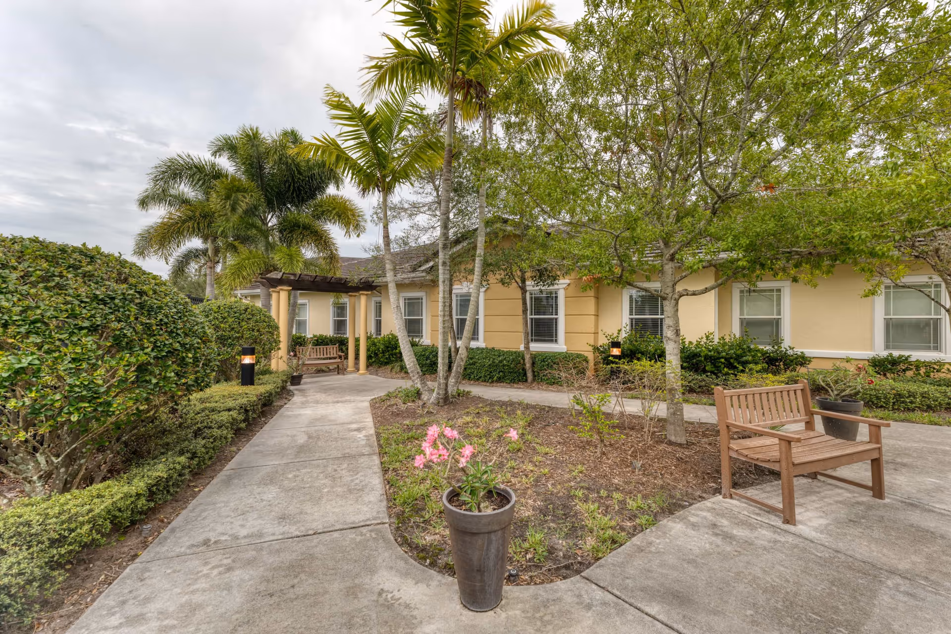 Outdoor garden area at Addington Place of Stuart featuring a concrete walkway, trimmed bushes, palm trees, a wooden bench, and a large planter with pink flowers in front of a beige building with white-trimmed windows.