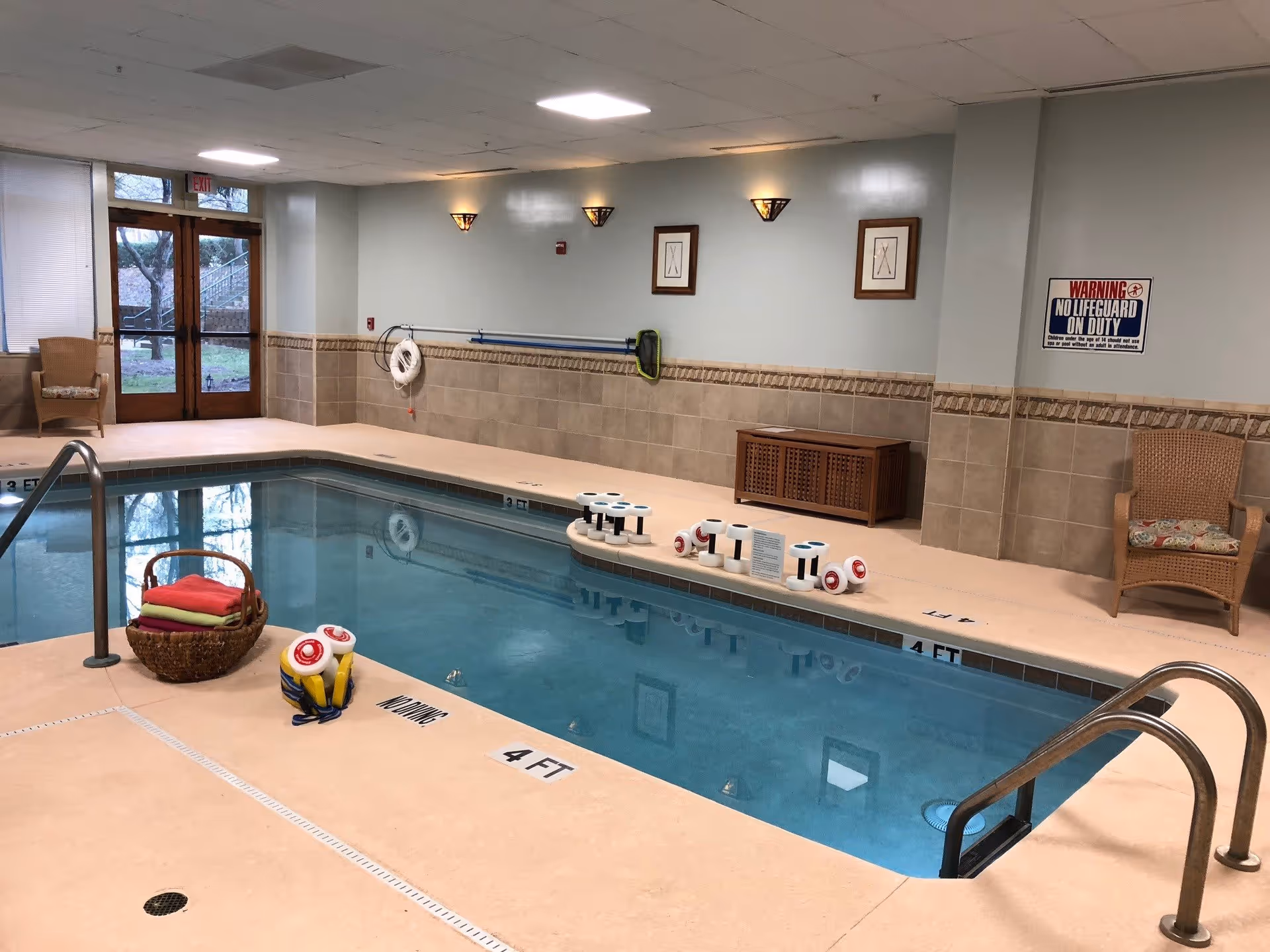 Indoor swimming pool area with handrails, pool toys and equipment, tiled walls, and wicker seating.