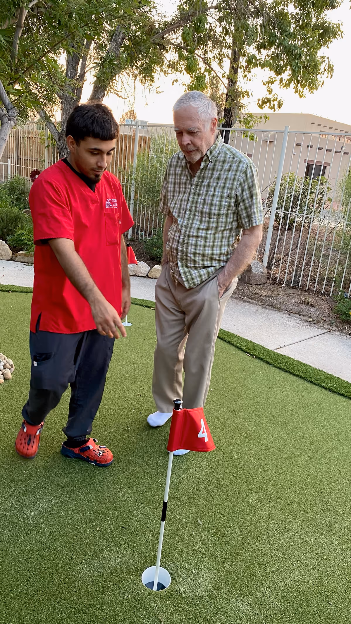 An elderly man and a younger man in a red shirt stand on a putting green outdoors. The younger man is pointing towards the hole marked with a red flag numbered 4, while the elderly man watches attentively. They are surrounded by a fenced garden area with trees and shrubs.