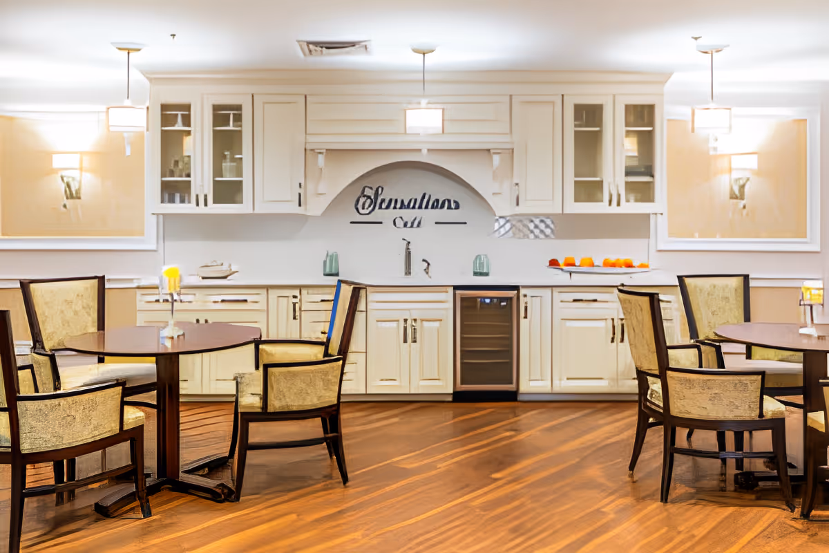 A well-lit dining area with two round wooden tables, each surrounded by four upholstered chairs. Behind the tables is a white cabinetry setup with glass-front upper cabinets, a small sink, and a wine cooler. The wall above the sink features the words 'Sensation Bar' in decorative script. The room has wooden flooring and soft wall lighting.