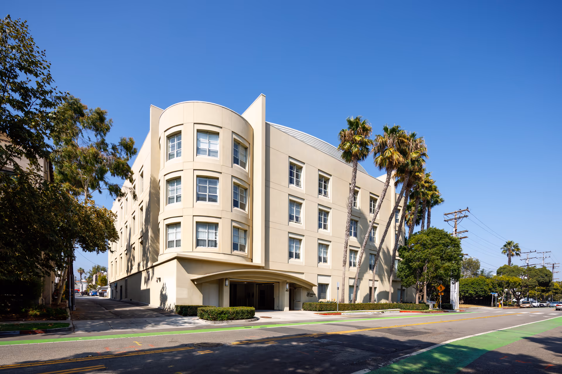 Exterior view of a multi-story beige building with rounded corner windows and palm trees lining the sidewalk under a clear blue sky.