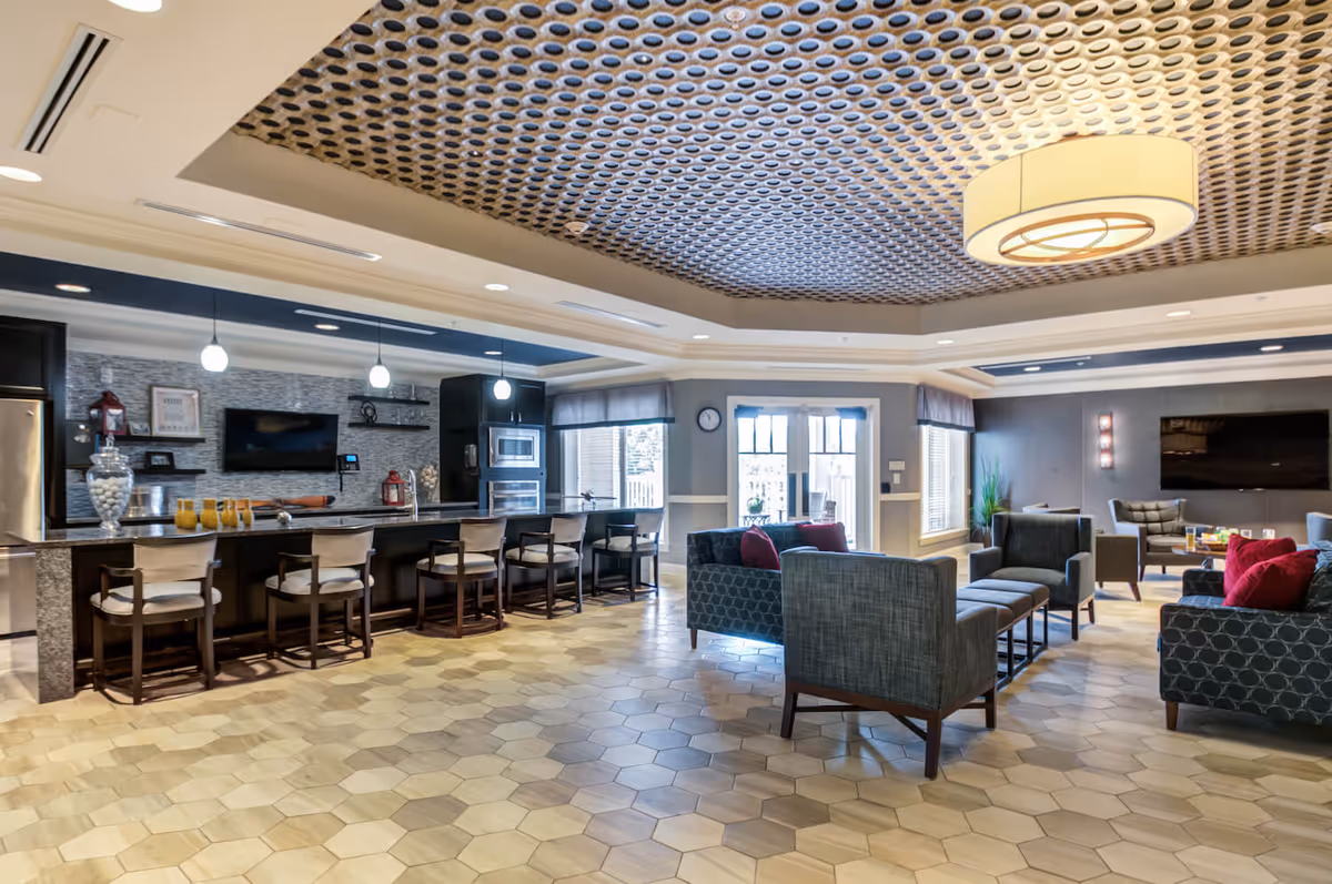 A spacious and modern common area in a senior living facility featuring a kitchen with a long counter and bar stools, a seating area with armchairs and sofas with red cushions, a large flat-screen TV mounted on the wall, and a decorative ceiling with circular patterns. The room has large windows and glass doors allowing natural light to enter.