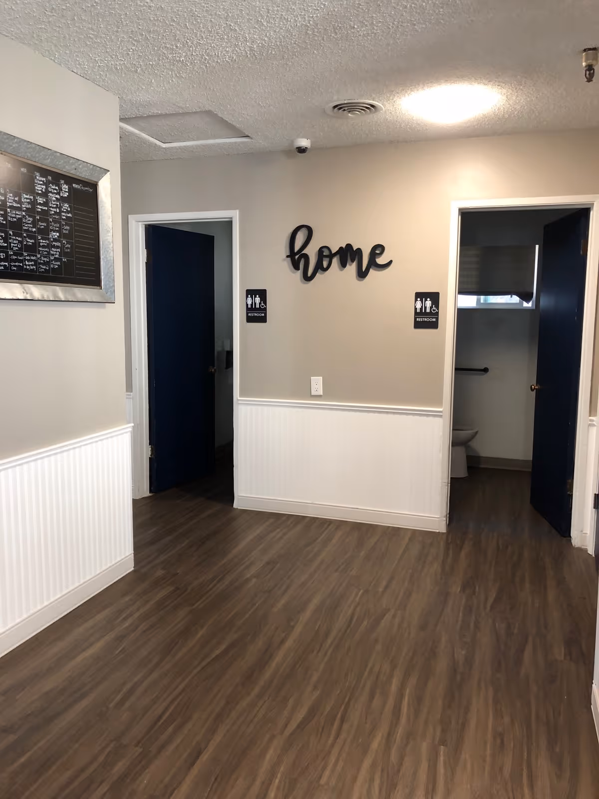 Interior hallway with wood-look flooring and light gray walls with white wainscoting. Two open doors with dark blue interiors lead to restrooms, indicated by black and white restroom signs with accessibility symbols. Above the restrooms is a decorative black script word 'home' mounted on the wall. A framed board is partially visible on the left wall.