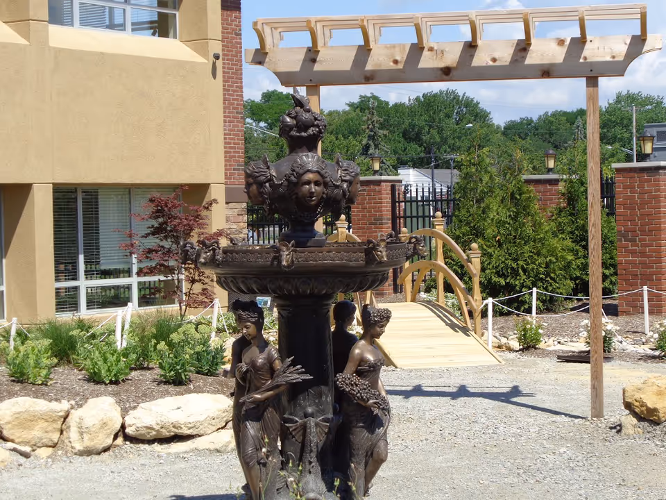 Outdoor garden area at The Carlyle House featuring a decorative bronze fountain with sculpted figures, a wooden pergola, a small arched wooden bridge, and surrounding greenery and landscaping with a building in the background.
