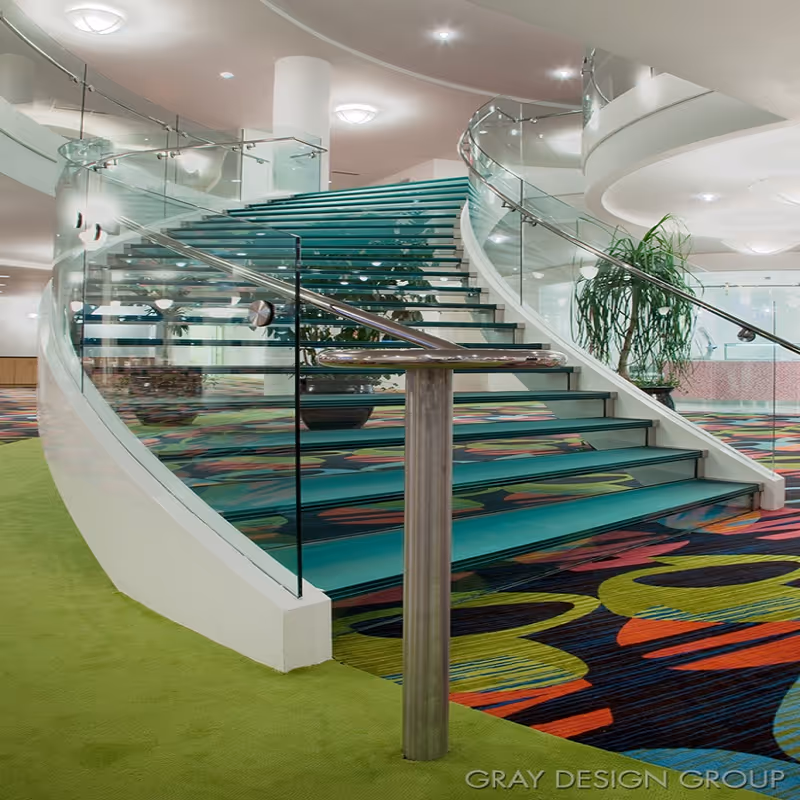 Curved staircase with glass railings and metal handrails in a modern interior space with colorful patterned carpet and green flooring, along with potted plants and ceiling lights.