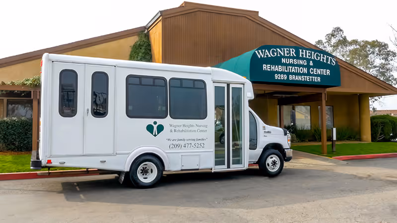 A white shuttle bus is parked in front of the Wagner Heights Nursing & Rehabilitation Center entrance with a green awning displaying the facility name.