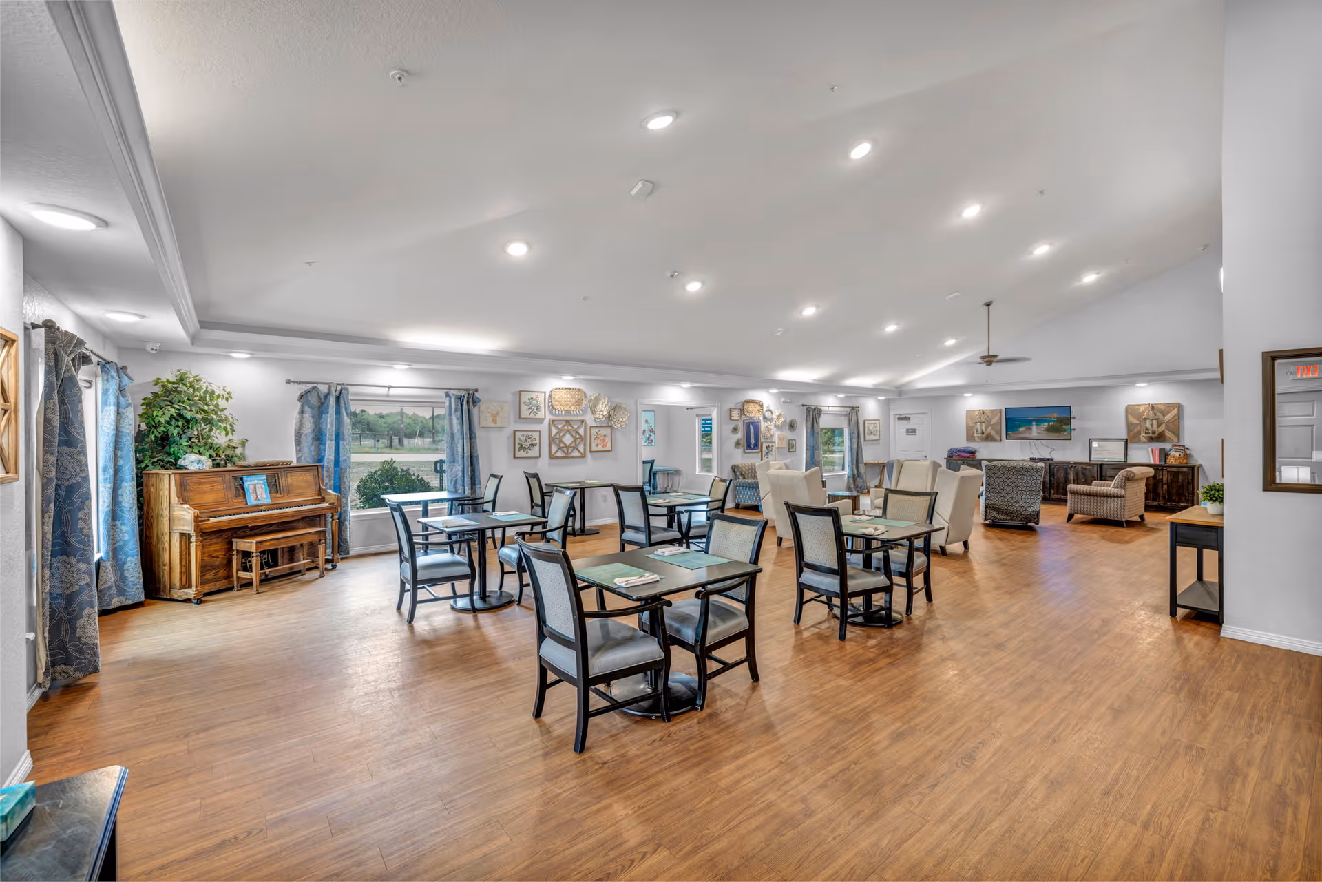 Spacious senior living community dining and lounge area with multiple tables and chairs, a piano by the window, and seating grouped near a TV.