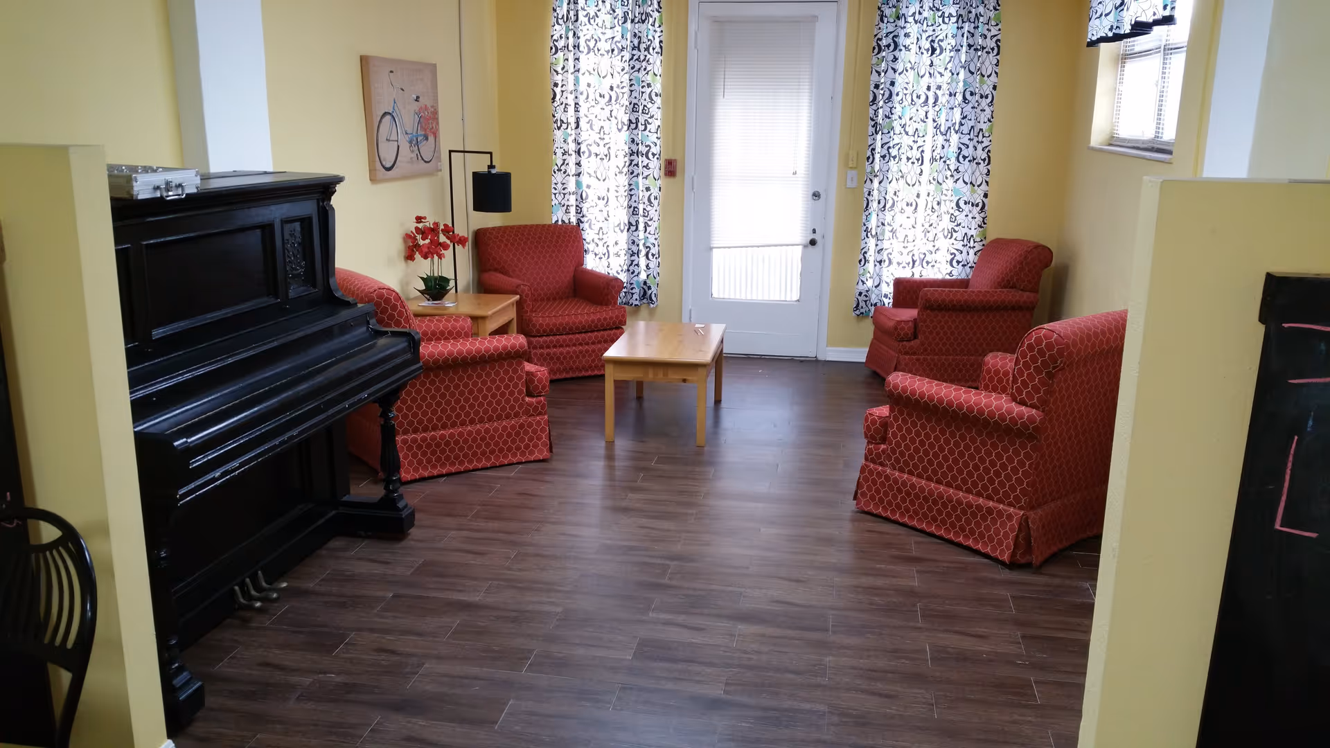 A cozy living room with four red patterned armchairs arranged around two wooden coffee tables. There is a black upright piano on the left side, yellow walls, and two windows with patterned curtains flanking a white door with blinds. A floor lamp and a wall painting of a bicycle are also visible.