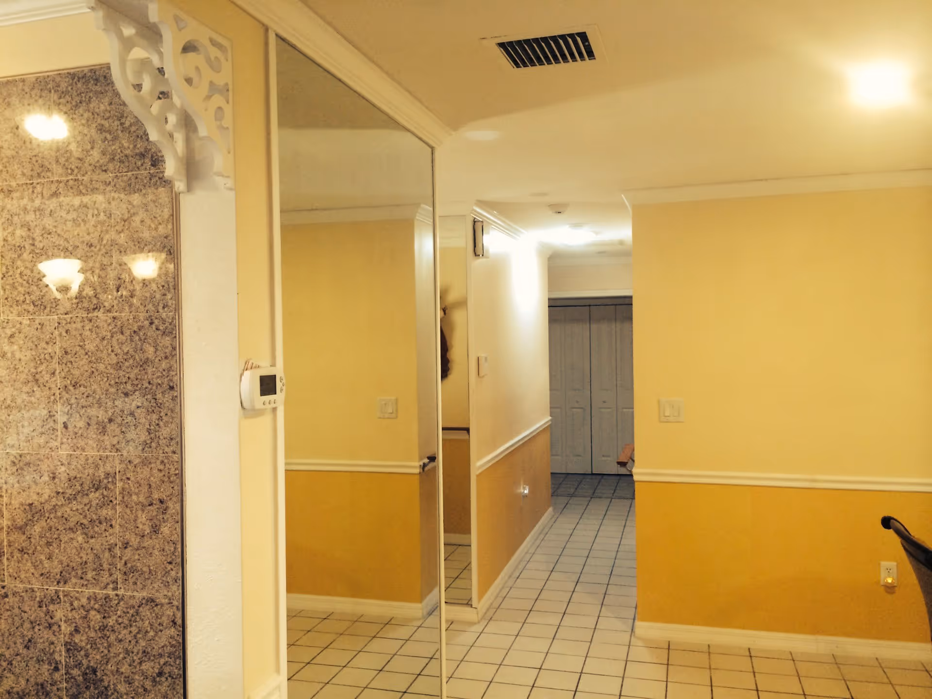 Interior hallway with yellow walls and white trim, tiled floor, a large mirror on the left wall, and a closed double door at the end of the hallway. A thermostat is mounted on the left wall near the mirror, and a portion of a granite wall is visible on the far left.
