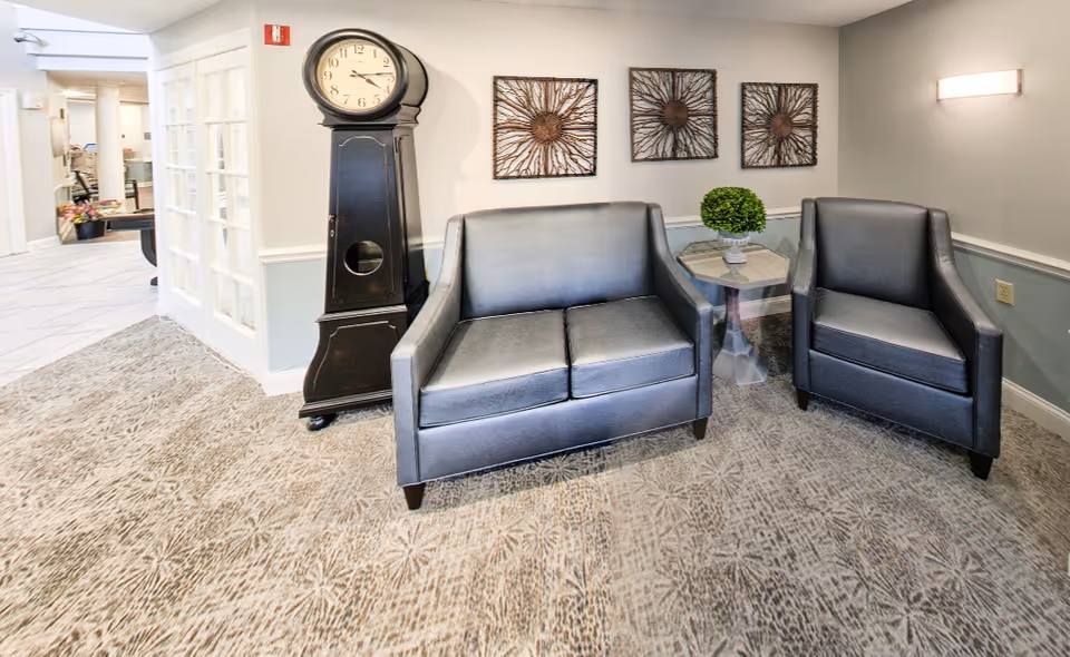 A cozy seating area in a senior living facility with a black leather loveseat and matching armchair. Between them is a small hexagonal side table with a decorative green plant. Behind the loveseat is a wall adorned with three metal sunburst wall art pieces. To the left of the loveseat stands a tall black grandfather clock. The room has patterned carpet flooring and light-colored walls with a chair rail. A hallway with white tiled flooring and glass-paneled double doors is visible in the background.
