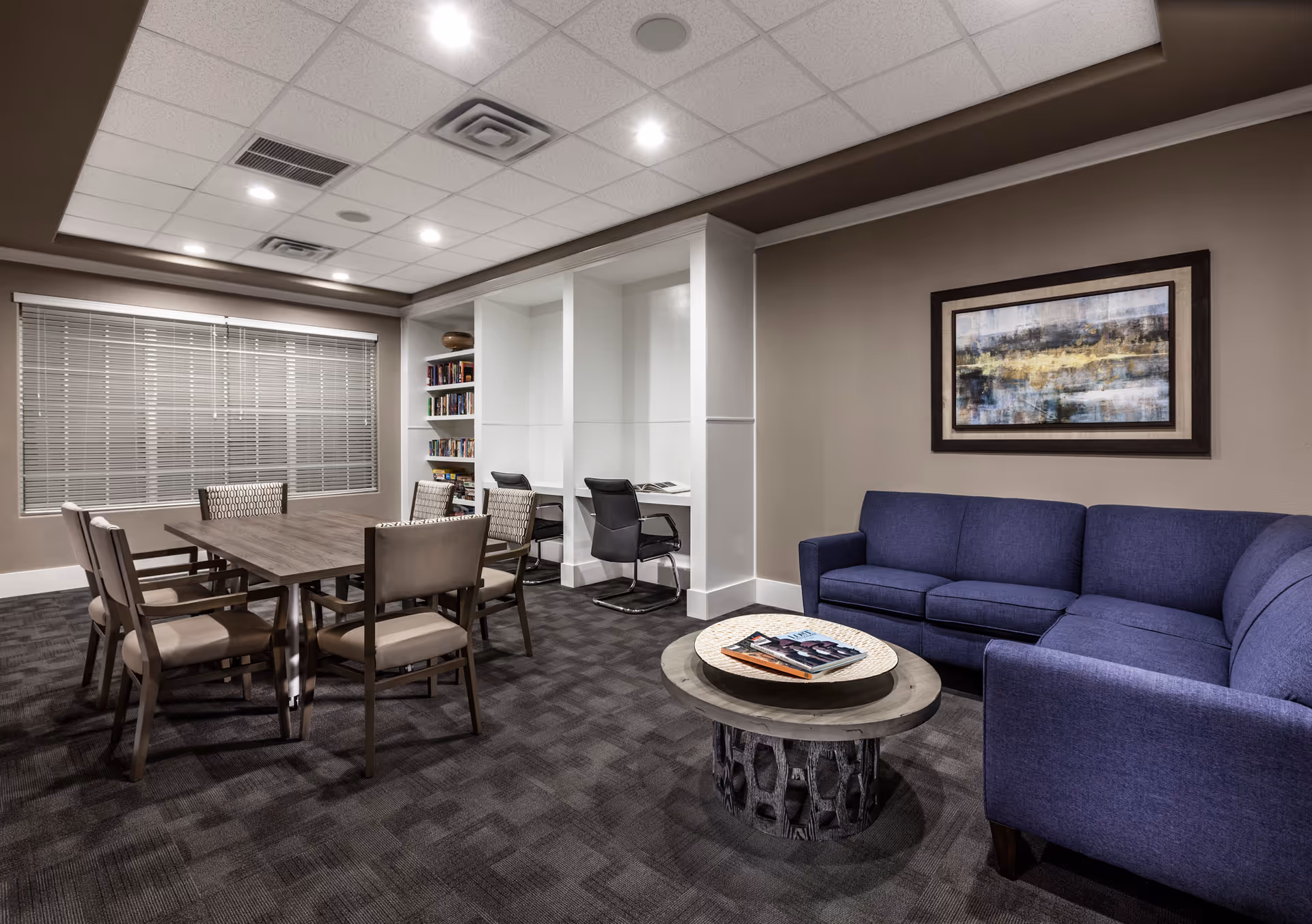 A well-lit interior room featuring a blue sectional sofa with a round wooden coffee table in front, holding magazines. To the left, there is a rectangular wooden table surrounded by six chairs. The back wall has built-in white shelves and two desk spaces with black chairs. A large window with closed blinds is on the left wall, and a framed abstract painting hangs above the sofa.