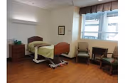 A simple, clean bedroom in a senior living facility with a single hospital-style bed with green bedding, a wooden nightstand with a phone and tissues, two armchairs, a small wooden table, and a large window with blinds letting in natural light.