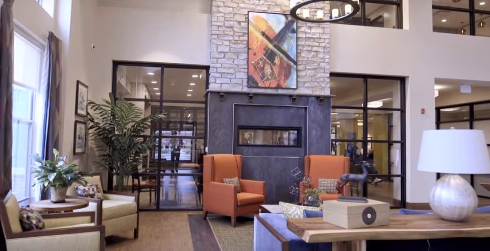A cozy and modern living room area with two orange armchairs facing a black fireplace set in a stone wall. There are large windows on the left side with curtains, a tall green plant, and a wooden table with a lamp and decorative items. The room has a high ceiling and glass doors leading to another area.