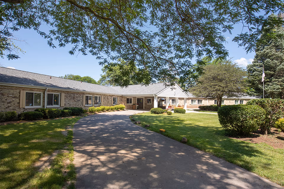 Exterior view of Madison Health and Rehabilitation Center showing a single-story brick building with multiple windows, a paved driveway leading to the entrance, well-maintained green lawns, bushes, and trees under a clear blue sky.