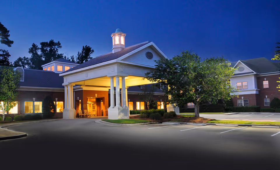 Exterior view of Laurel Crest Retirement Community building at dusk with lights glowing from windows and a covered entrance with columns. There is a parking lot in front with some trees and landscaping.