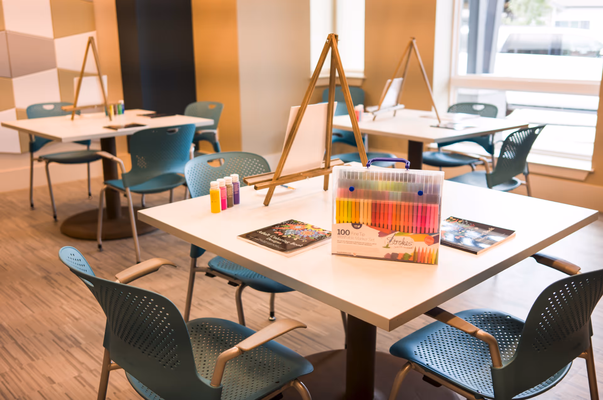 A bright activity room with several tables and teal chairs arranged around them. Each table has a wooden easel, paint bottles, coloring books, and a set of colorful markers. Large windows allow natural light to fill the space.