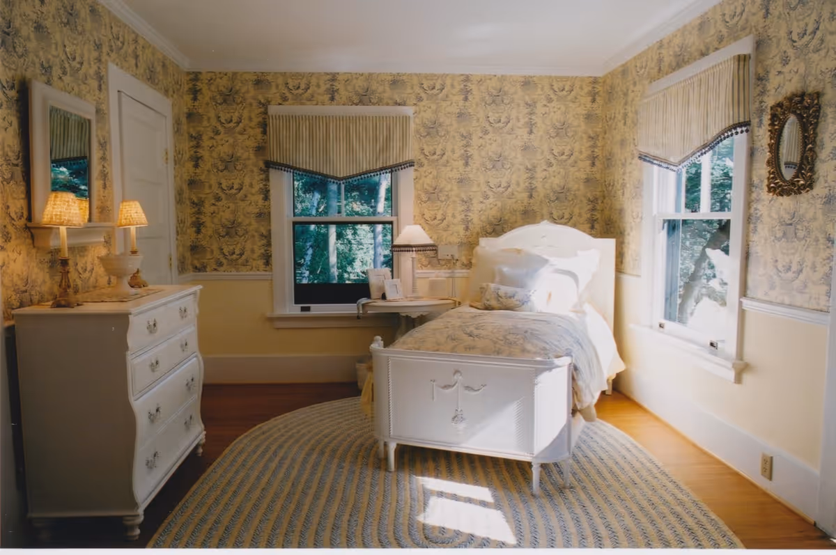 A cozy bedroom with vintage-style decor featuring a white wooden bed with floral bedding, a white dresser with two lit lamps and a mirror above it, two windows with striped valances letting in natural light, and a round braided rug on a wooden floor. The walls have yellow floral wallpaper with a white chair rail.