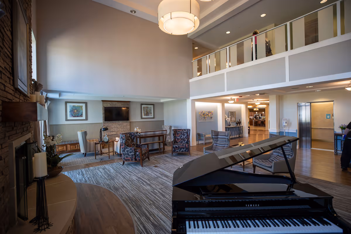 Spacious senior living facility common area with a grand piano in the foreground, comfortable seating including patterned armchairs and high-back chairs, a fireplace with candles, framed artwork on the walls, a flat-screen TV mounted above a stone accent wall, and a second-floor balcony overlooking the room.