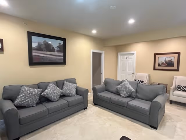 A cozy living room with two gray sofas each adorned with patterned throw pillows. The walls are painted a soft beige and decorated with framed artwork. There are two white armchairs with patterned cushions in the corner near a closed white door. The room is well-lit with recessed ceiling lights and has a light-colored carpeted floor.
