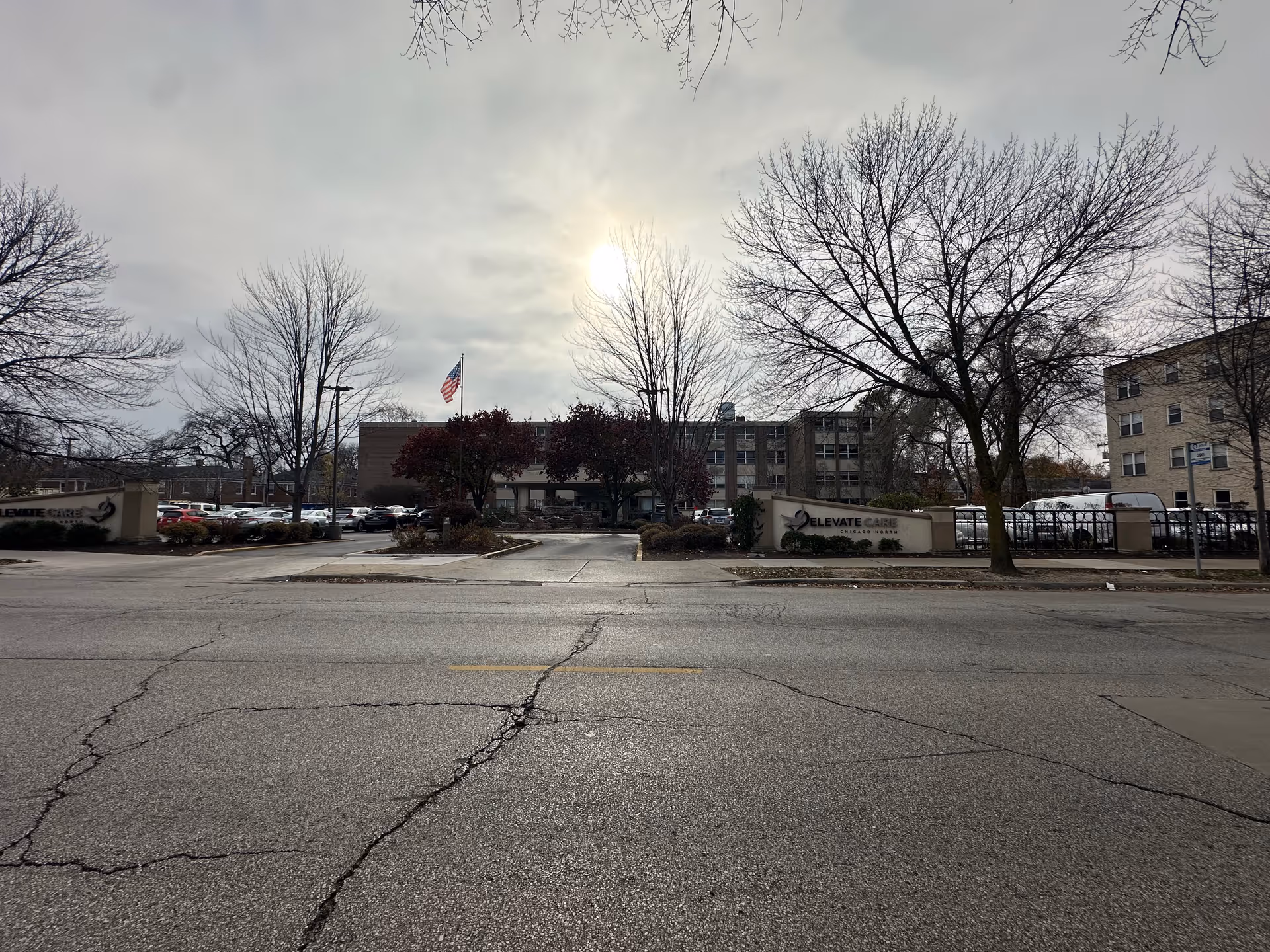 Exterior view of Elevate Care Chicago North facility with a parking lot, leafless trees, and an American flag flying on a flagpole under a cloudy sky.