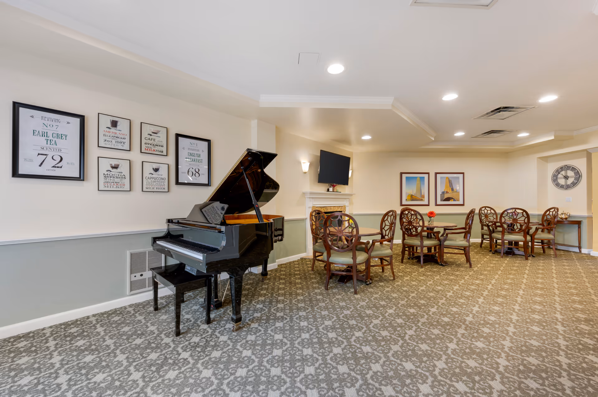 A spacious common area in a senior living facility featuring a black grand piano, several round wooden tables with chairs, framed artwork on the walls, a wall-mounted TV above a fireplace, and a large wall clock. The room has patterned carpet flooring and recessed ceiling lights.