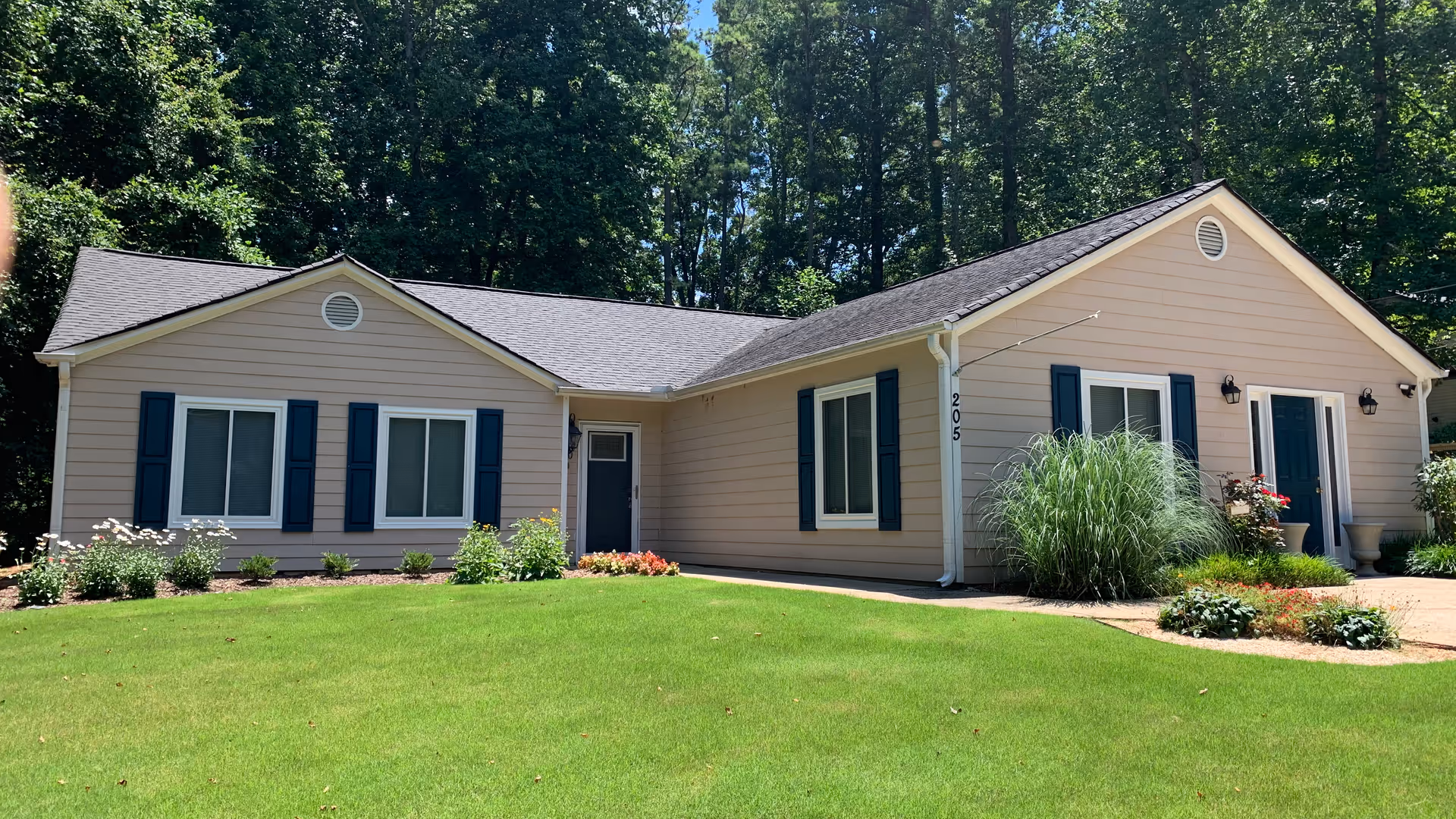 Single-story beige house with blue shutters, a green lawn and landscaped plants in front of a wooded backdrop.