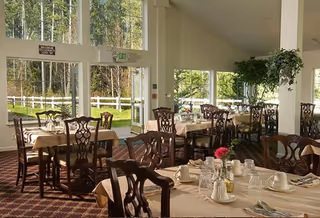 Dining room with multiple tables covered in beige tablecloths, set with cups, glasses, and silverware. Wooden chairs with intricate backs surround the tables. Large windows allow natural light to fill the room and provide a view of a green outdoor area with trees and a white fence.