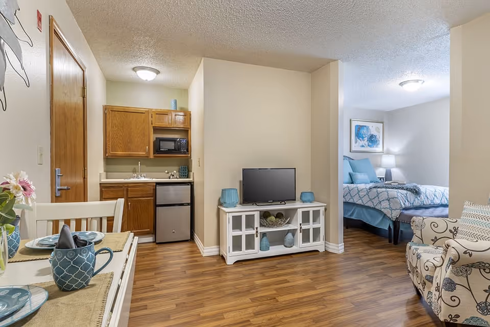 Interior view of a senior living facility apartment showing a small kitchenette with wooden cabinets, a microwave, and a mini refrigerator. To the right is a white TV stand with a flat-screen TV and decorative items. Further right is a cozy bedroom area with a bed dressed in blue and white bedding, a nightstand with a lamp, and a framed artwork on the wall. In the foreground, there is a dining table set with blue dishware and a floral centerpiece, and a patterned armchair is partially visible on the right. The flooring is wood laminate throughout.