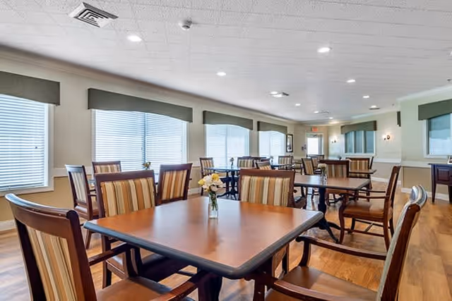 A bright dining room with multiple wooden tables and chairs arranged neatly. Each table has a small vase with flowers. Large windows with blinds allow natural light to fill the room. The walls are light-colored with green valances above the windows and wooden flooring.