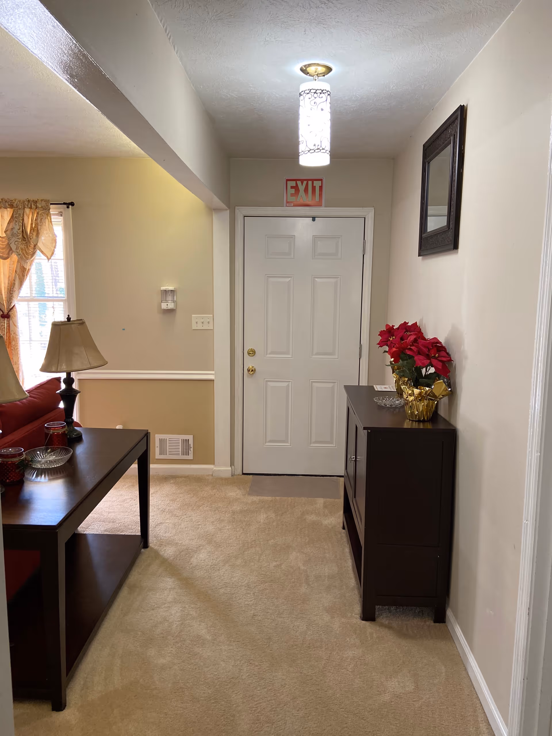Interior view of an entryway showing a white door with an EXIT sign above, a console table with a poinsettia, and a nearby lamp and sofa.