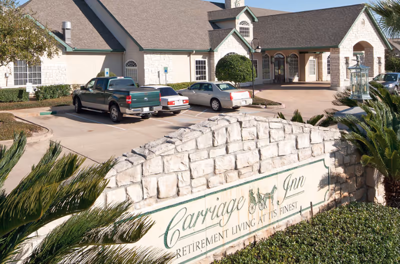 Exterior view of Carriage Inn Lake Jackson retirement living facility showing a stone sign with the facility name and slogan, a parking lot with several vehicles, and a building with a covered entrance and arched windows.