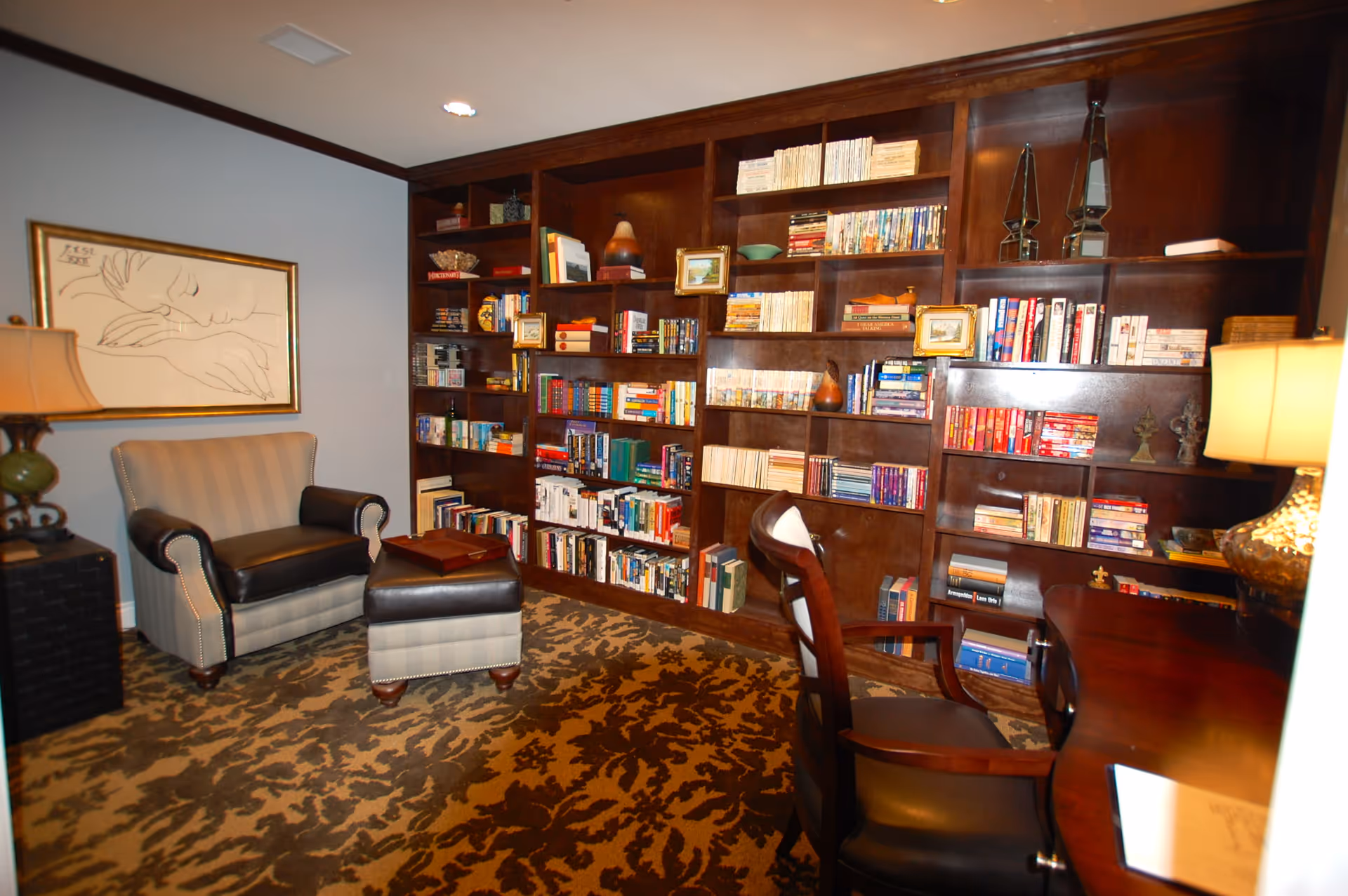A cozy library room with a large dark wooden bookshelf filled with books and decorative items. There is a striped armchair with a matching ottoman, a wooden desk with a chair, and two table lamps providing warm lighting. A framed line art drawing hangs on the light blue wall.