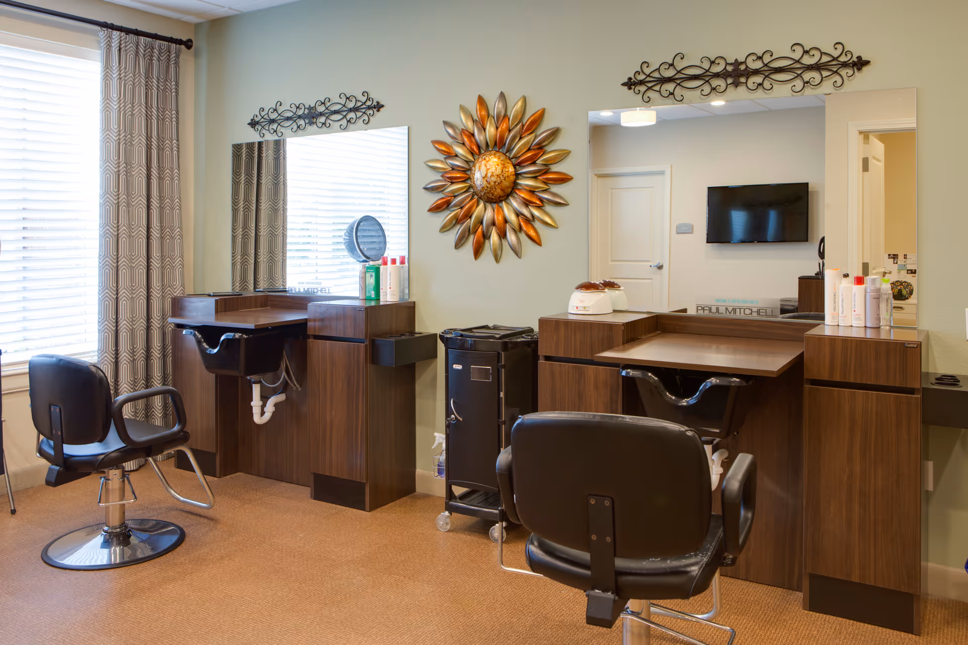 Interior view of a hair salon area with two styling stations, each equipped with a large mirror, a black salon chair, and wooden cabinetry. The room has a window with patterned curtains, a decorative sunburst wall art, and various hair care products on the counters.