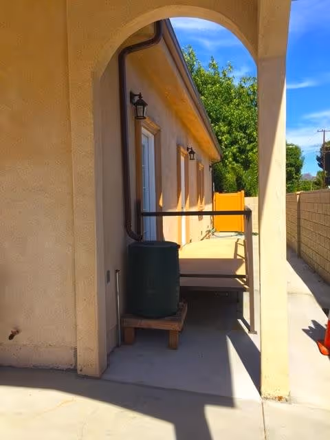 Outdoor view of a beige stucco building with an arched walkway. There is a green rain barrel on a wooden pallet next to the building, a small raised wooden deck with a metal railing, and a yellow gate at the end of the walkway. Trees and a blue sky are visible in the background.