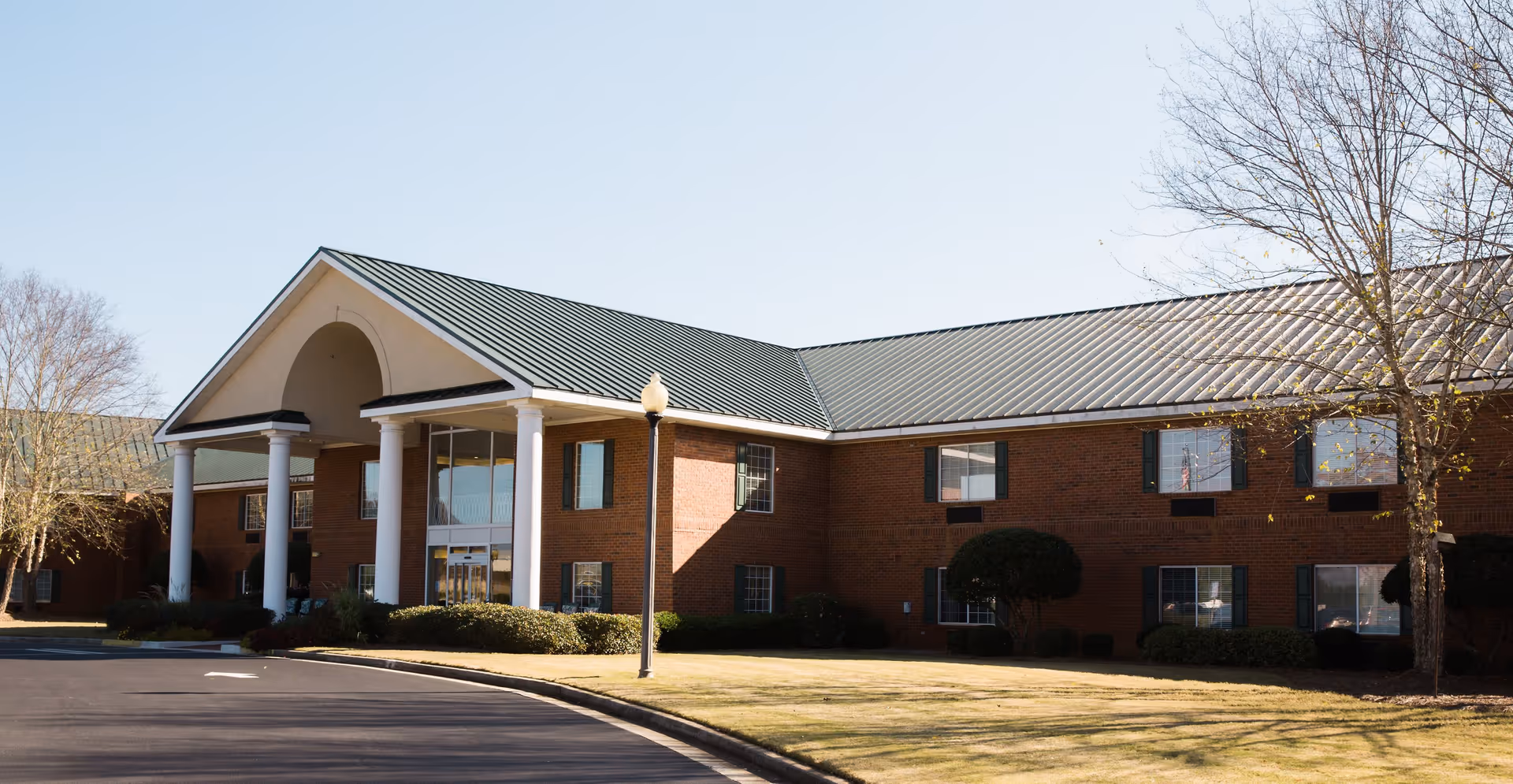 Exterior view of a two-story senior living facility building with a brick facade, green metal roof, white columns at the entrance, and a curved driveway in front. There are some leafless trees and bushes around the building.
