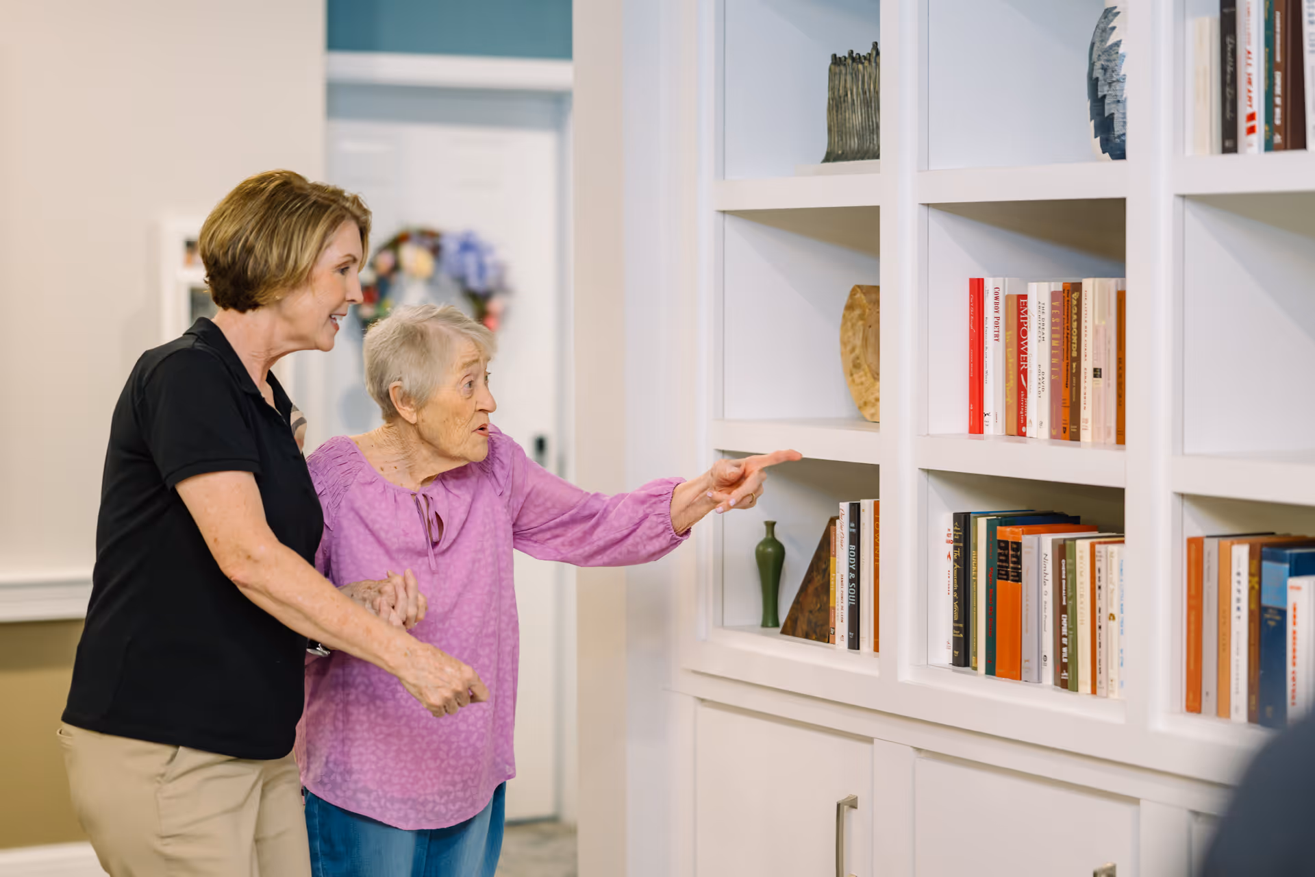 An elderly woman in a purple blouse is pointing at a white bookshelf filled with books and decorative items, while a caregiver in a black shirt and beige pants supports her from behind in a well-lit room.