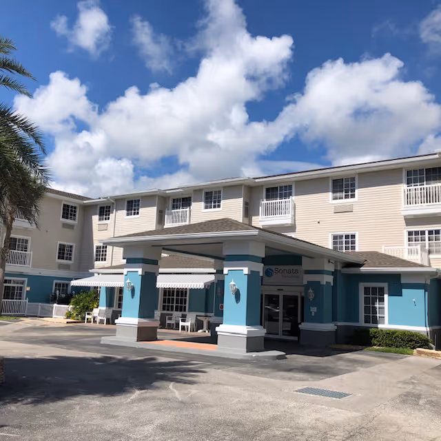 Front exterior of a three-story Sonata by AgeWell Solvere Living building with a covered entrance and blue accents under a partly cloudy sky.