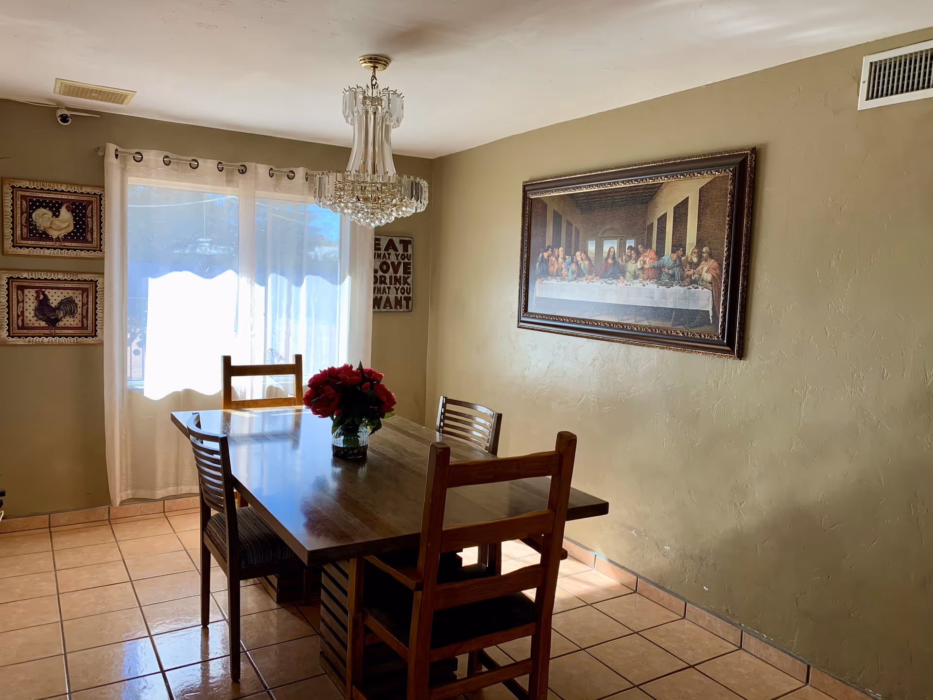 Sunlit dining room with a wooden table and chairs, a chandelier, a vase of red flowers, and framed artwork on the walls.