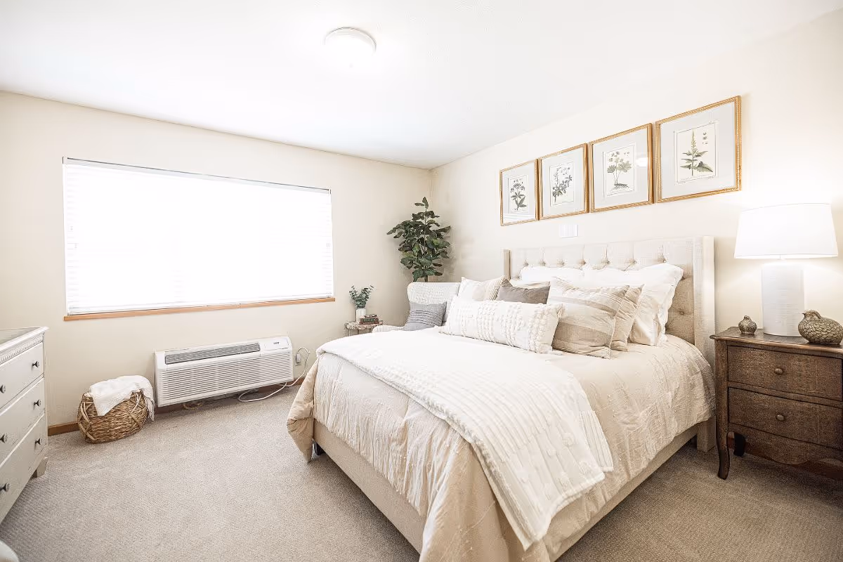 Bright, neutral bedroom with a made bed, bedside tables and lamp, framed botanical prints, and a large window.