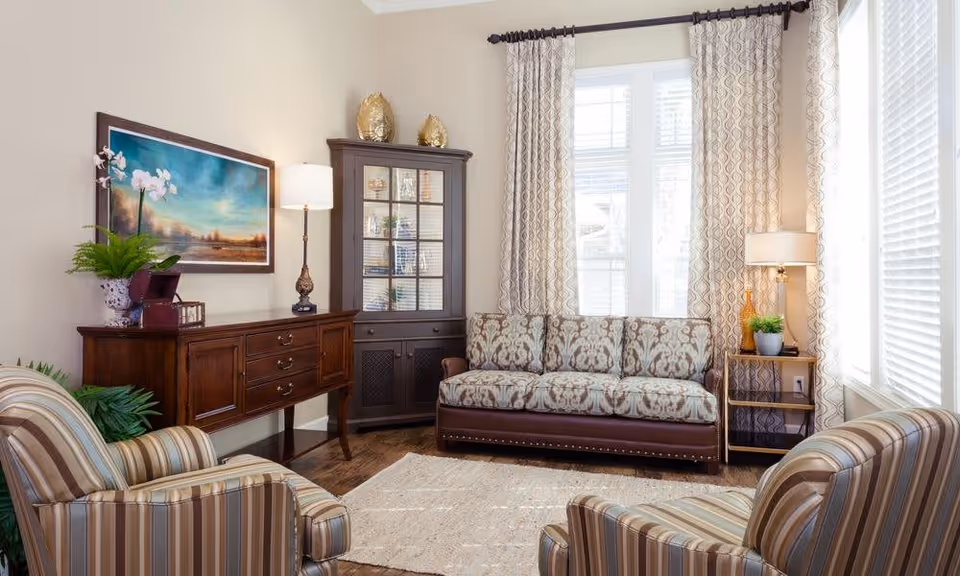 A cozy living room with a patterned three-seater sofa, two striped armchairs, a wooden sideboard with a lamp and decorative items, a glass-front cabinet, a floor lamp on a small side table, and large windows with patterned curtains letting in natural light.