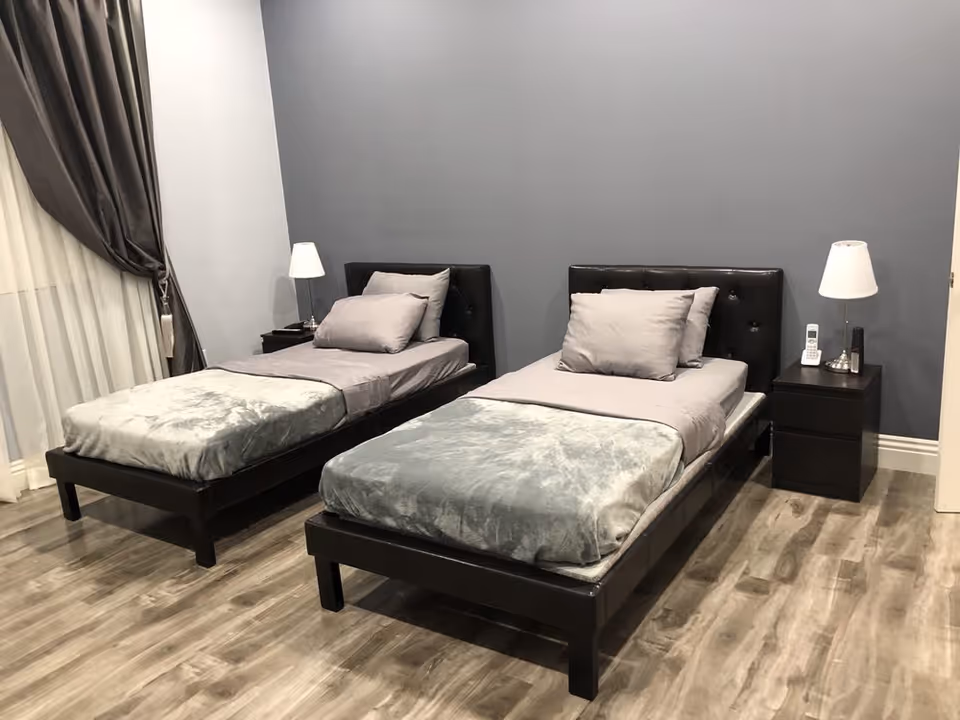 A bedroom with two single beds featuring black headboards, gray bedding, and pillows. Each bed has a black nightstand beside it with a white lamp. The room has wooden flooring, gray walls, and a window with dark curtains on the left side.